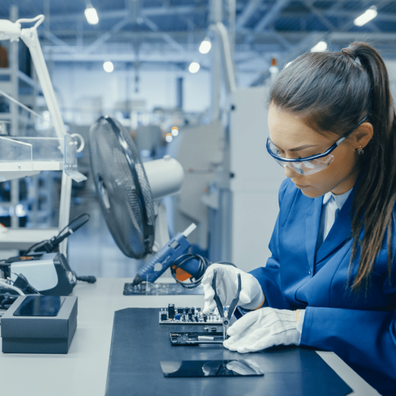 Opératrice en blouse et lunettes de protection assemblant une carte électronique sur un poste d’intégration en atelier