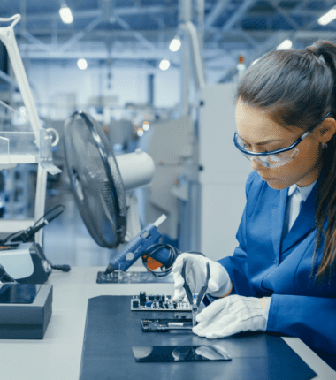 Technicienne en laboratoire industriel assemblant une carte électronique avec des outils de précision, portant des gants et des lunettes de protection